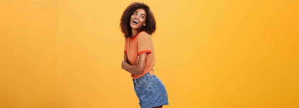 Stock image Hey wanna hang out. Sensual and flirty carefree stylish dark-skinned woman with curly hair standing in profile turning left and posing with happy cute expression smiling and seducing over orange wall.