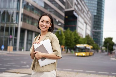 Güzel Asyalı kız işe giderken gülümsüyor, dizüstü bilgisayar ve defterle sokakta duruyor..