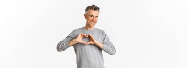 Yüzsüz orta yaşlı bir adamın portresi gri kazaklı, gülümseyen ve kalp işareti gösteren, beyaz arka planda duran birini seven.