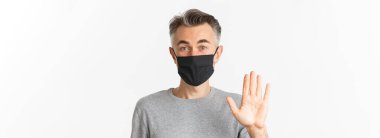 Covid-19 kavramı, sosyal uzaklık ve karantina. Siyah tıbbi maskeli, dost canlısı orta yaşlı adamın yakın çekimi. Merhaba deyip el sallıyor, birini selamlıyor, beyaz arka planda duruyor..