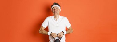Egzersiz anlayışı, spor salonu ve yaşam tarzı. Çaydanlığı kaldırmaya çalışan komik orta yaşlı sporcunun portresi, turuncu arka planda duruyor..