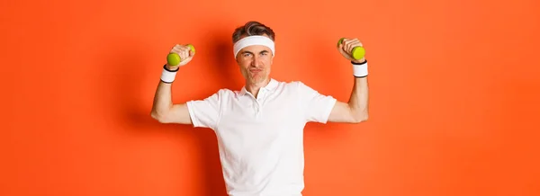 stock image Portrait of cheeky middle-aged fitness guy, doing sports against orange background, flex biceps and holding dumbbells.