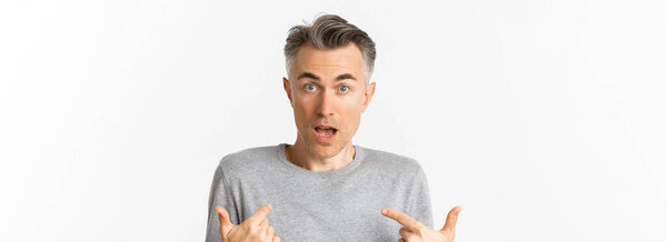 Close-up of confused middle-aged man, pointing at himself and gasping, standing over white background.