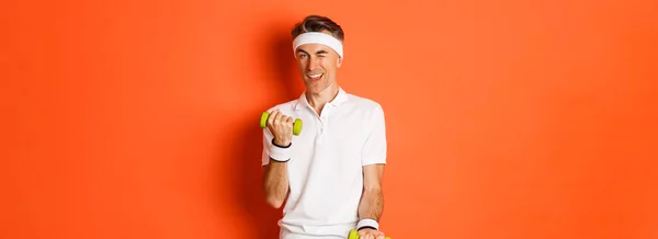 厚脸皮 中年男性运动员参加体育运动 带着哑铃运动 在镜头前眨眼 站在橙色背景上的画像 — 图库照片