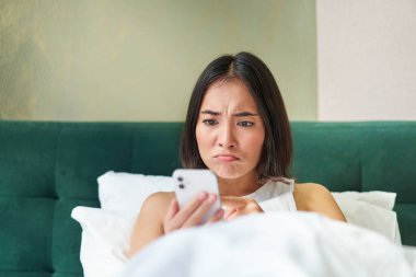 Huysuz Asyalı kızın yakın portresi. Yatakta uzanıyor, cep telefonunda karmaşık bir konu yüzünden surat asıyor ve surat asıyor. Akıllı telefon uygulaması okuyor..