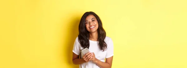 美しいアフリカ系アメリカ人女性の肖像 笑顔とスマートフォンを保持 黄色の背景の上に立って — ストック写真