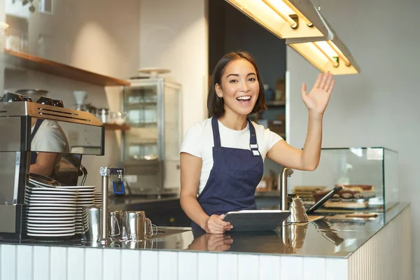 陽気なアジアの少女バリスタカウンターの後ろのバーテンダー腕を上げ 顧客に手を振って カフェで注文を処理し コーヒーショップで働く — ストック写真