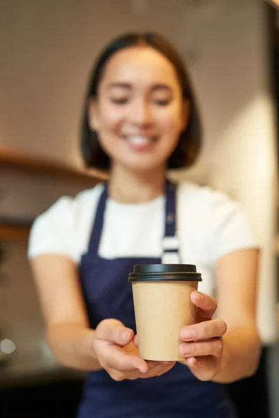 笑顔アジアの少女バリスタは クライアントに注文を与えます テイクアウトコーヒーカップを保持 エプロンを身に着けています カフェでカウンターの後ろに働く — ストック写真