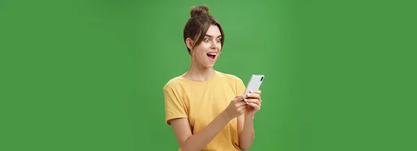 女人在智能手机上看到令人惊叹的令人满意的信息时 兴奋得张开嘴 笑着惊讶地看着手机屏幕在绿色背景下穿着休闲的黄色T恤 — 图库照片