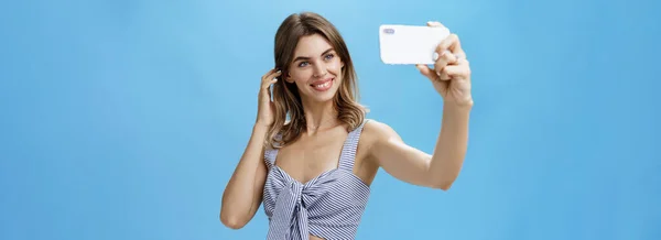スマートフォンの画面で楽しそうに笑顔耳の後ろに髪をフリックスタイリッシュなマッチング衣装で優れた自尊心を持つ魅力的な女性 インターネットに投稿し 青い壁にフォロワーを得るために自撮りを取ります — ストック写真