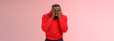 Üzgün, Afro-Amerikan sakallı, kırmızı kapüşonlu sakallı adam yüzünü parmaklarının arasından gizliyor üzgün, harap olmuş, üzgün, pembe arka planda duran kederli, üzgün, kederli.