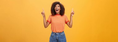 Afro saçlı, şık tişörtlü ve kot şortlu, turuncu duvardan yukarı bakan, etkileyici ve eğlenceli Afro-Amerikan kadın.