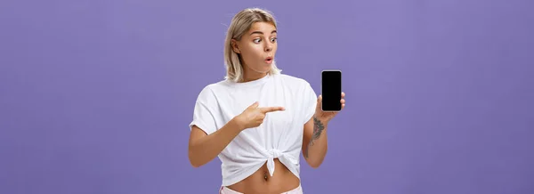 ブロンドの髪と入れ墨が紫色の背景の上に立って素晴らしいブランドの新しいスマートフォンを見て 指している印象的で喜んで豪華なヨーロッパの女性のウエストアップショット 技術コンセプト — ストック写真