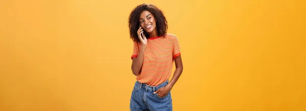 Stylish dark-skinned girl making casual phone call to friend telling all details of after romantic date standing pleased and carefree over orange background in striped t-shirt gazing left with grin