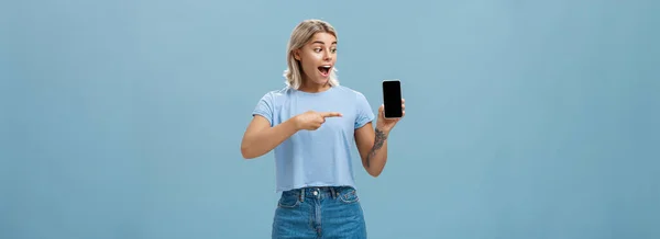 この素晴らしいスマートフォンを見てください 熱いです良いです見た目の良いです女性ブロンドとともにクールな入れ墨アームドロップ顎からスリルと興奮ポインティングでデバイス画面上の青の背景 — ストック写真