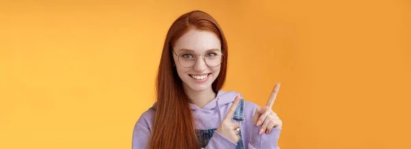 自信的 漂亮的 面带微笑的红头发的自由职业女子 戴着眼镜 指着左上角食指 咧嘴笑着 牙齿洁白 自信地提出建议 应该是防弹的 橙色的背景 — 图库照片