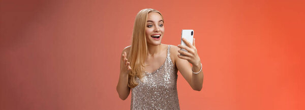 Charming elegant nice blond girl in silver dress talking video call speaking looking smartphone display amused surprised smiling happily have conversation sibling showing prom outfit.