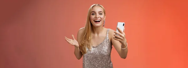 stock image Astonished happy charming blond girl in silver glittering stylish dress holding smartphone amazed liking awesome result edit photo app smiling wondered amused, standing red background.