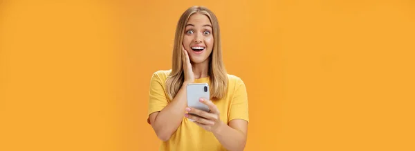令人惊讶和印象深刻的女性形象 在智能手机中对令人敬畏的应用程序做出了反应 带着惊讶和喜悦的表情面对手持手机翻过橙色墙壁 — 图库照片