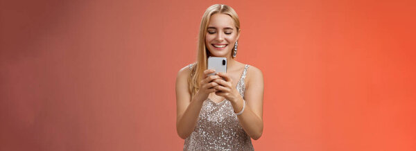 Delighted tender glamour blond woman in silver stylish glittering dress brilliand earrings holding smartphone taking photo friend capture moment celebration nightclub, red background smiling.