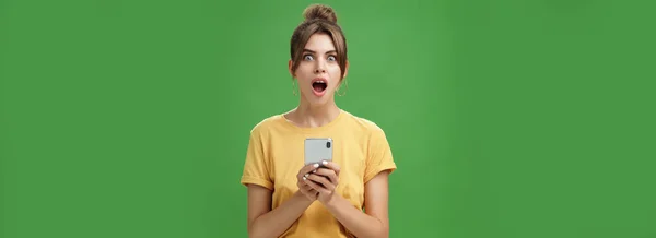ショックを受けた言葉と印象的な美しい白い女の子の肖像画黄色のTシャツを保持するスマートフォンで髪を櫛 緑の背景にクールなアプリに反応興奮から顎をドロップ — ストック写真