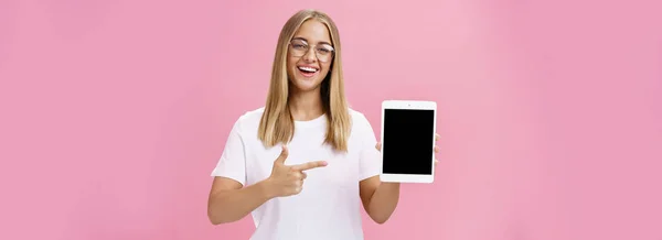 女性自由职业程序员自豪地展示了她的数字平板电脑应用程序 手持小玩艺儿指着屏幕 带着喜庆的表情 戴着眼镜 在粉红的墙上摆出一副笑脸 — 图库照片