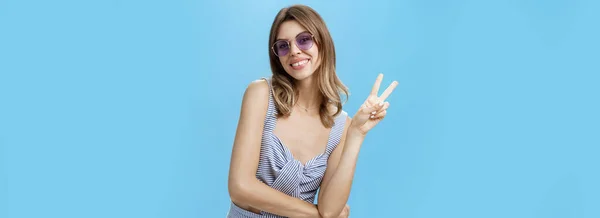 stock image Charming friendly and outgoing young lady showing peace or victory gesture and smiling broadly at camera showing cute gapped teeth, tilting head and raising one shoulder posing flirty over blue wall.