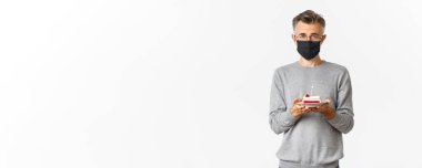 Koronavirüs, karantina ve tatil kavramı. Siyah tıbbi maskeli ve gözlüklü, elinde mumla doğum günü pastası tutan üzgün orta yaşlı bir adam imajı..