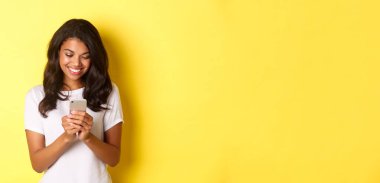 Beyaz tişörtlü Afro-Amerikalı güzel bir kızın portresi, akıllı telefonuyla mesajlaşıyor, cep telefonu uygulaması kullanıyor, sarı arka planda duruyor..