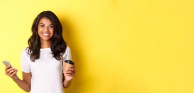 Çekici bir Afro-Amerikalı kızın portresi gülümsüyor, elinde kahve fincanı ve cep telefonu, sarı arka planda duruyor..