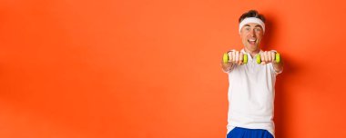 Aktif orta yaşlı fitness elemanının görüntüsü, dambıllarla spor egzersizleri yapıyor ve heyecanla gülümsüyor, turuncu arka planda duruyor..
