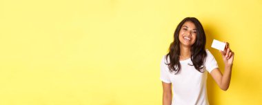 Afro-Amerikalı güzel bir kadının mutlu bir şekilde gülümsemesi, kredi kartı göstermesi, sarı arka planda durması..