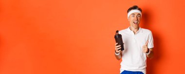 Sağlıklı ve aktif orta yaşlı fitness elemanı portresi, spor üniforması giyiyor, baş parmağını kaldırıyor, içme suyu içiyor, turuncu arka planda duruyor..