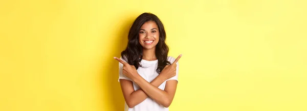 幸せと自信に満ちたアフリカ系アメリカ人の少女の肖像画は 右と左のコピースペースで横に指を指して 満足し 黄色の背景の上に立って — ストック写真