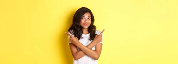 かわいいです優柔不断なアフリカ系アメリカ人の少女の肖像画 指を横に向けて 肩をすくめ 選択とアドバイスを求めます 黄色の背景に立って — ストック写真