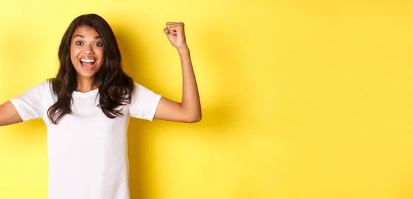 Stock image Image of happy african-american girl achieve goal and celebrating victory, raising hands up and smiling pleased, looking satisfied, standing over yellow background.