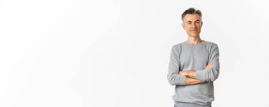 Çekici orta yaşlı adamın portresi elleri göğsünde çapraz duran, gülümseyen, beyaz arka planda poz veren.