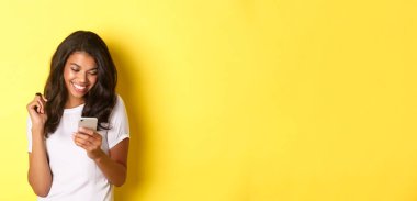 Beyaz tişörtlü, akıllı telefonuyla mesajlaşan, cep telefonuna bakıp gülümseyen, sarı arka planda duran çekici Afro-Amerikan kızı..