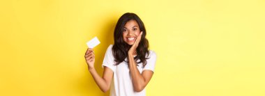 Afro-Amerikalı güzel bir kadının mutlu bir şekilde gülümsemesi, kredi kartı göstermesi, sarı arka planda durması..