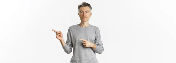 一个穿着灰色毛衣的有魅力的中年男子的画像 手指正对着 神色严肃 展示了一则广告 站在白色的背景之上 — 图库照片