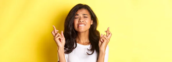 stock image Close-up of anxious african-american woman, pleading, biting lip and close eyes while making a wish, crossing fingers for good luck, standing over yellow background.