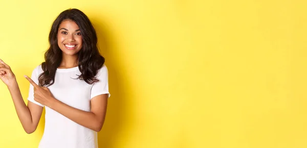 stock image Portrait of cheerful and cute african american girl, pointing fingers left and smiling, showing advertisement on copy space, standing over yellow background.
