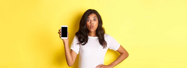 令人惊讶的非洲裔美国女孩的照片 她神魂颠倒地看着智能手机屏幕 站在黄色的背景上 — 图库照片
