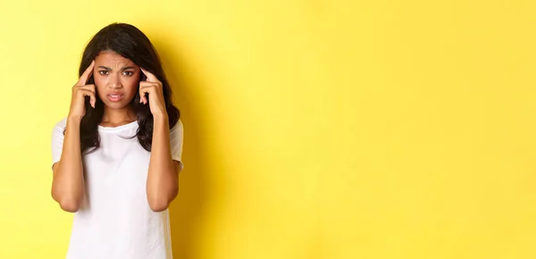 stock image Portrait of frustrated african-american girl, frowning and touching head, looking distressed at camera, standing over yellow background.