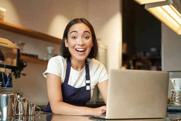 熱狂的なアジアの女の子でカフェ制服 ノートパソコンとバリスタ労働者 見て幸せと驚きでカメラ — ストック写真