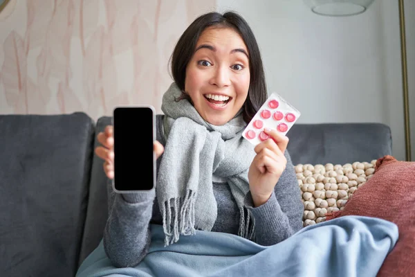 Online doctors and healthcare. Smiling korean woman shows mobile phone screen and medication, catching cold, staying at home and recommending smartphone GP.