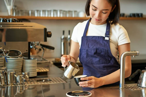 幸せな笑顔のカフェのオーナー エプロンの少女バリスタ カプチーノを作る 蒸しミルクとラテアート カウンターの後ろに立って — ストック写真