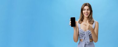 Karizmatik, şaşkın ve mutlu dişleri açık gülümseyen bir kadının portresi. Alet ekranını işaret eden harika bir akıllı telefonu tutarken şaşkın ve şaşkın bir şekilde alete bakıyor.