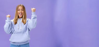 Neşeli, mutlu, destekleyici, açık renk saçlı, kapüşonlu, bronz tenli, neşeli ve neşeli genç bir kız arkadaş. Gülümseyen bir gülümsemeyle yumruklarını kaldırıyor.