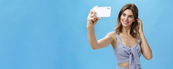 Attractive woman with good self-esteem in stylish matching outfit flicking hair behind ear smiling joyfully at smartphone screen, taking selfie to post in internet and gain followers over blue wall